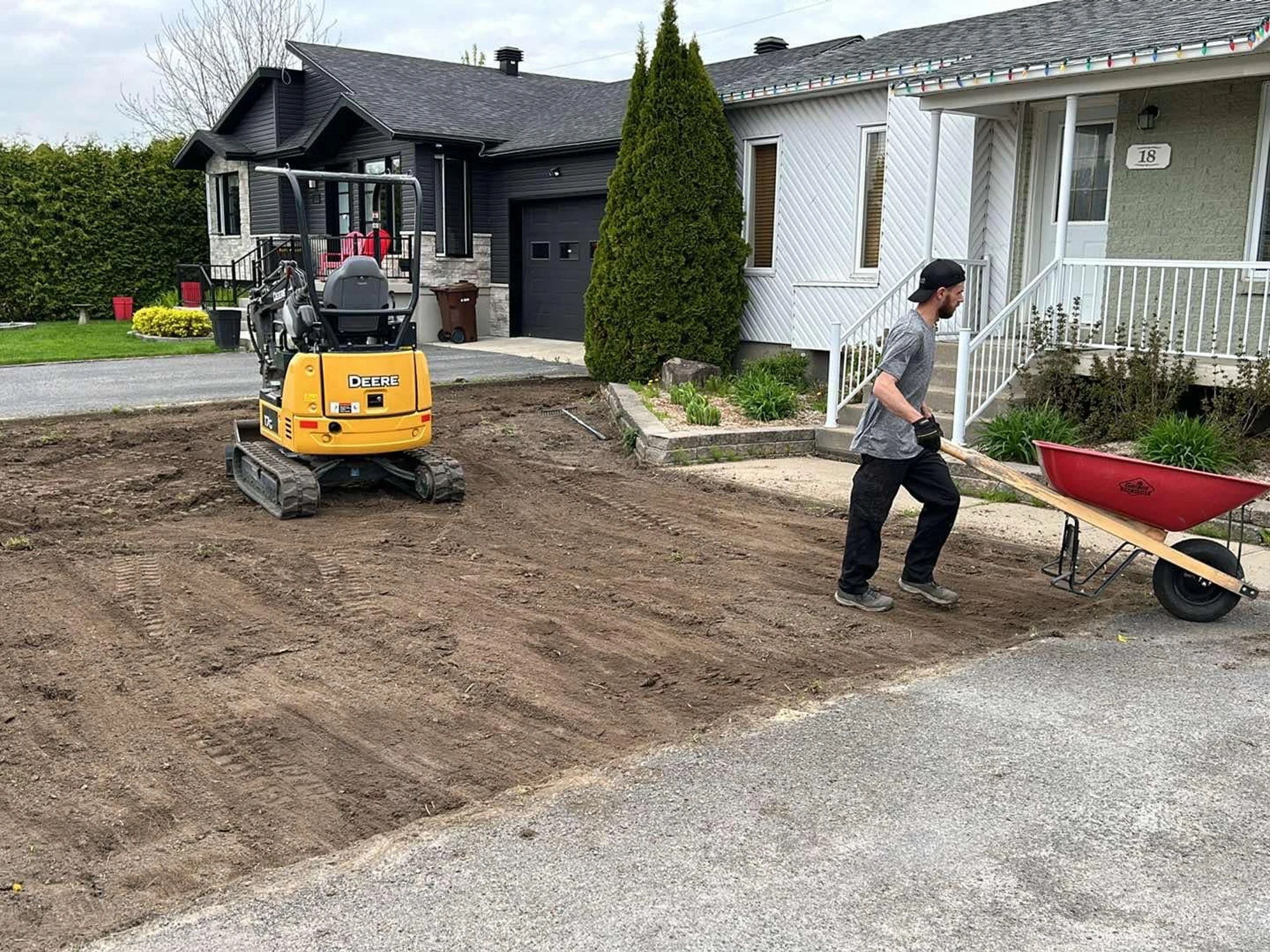 Mini-pelle en train de niveler un terrain devant une maison, ouvrier de TerraCare avec une brouette.